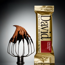 Fudge brownie flavored David protein bar next to a whisk with chocolate frosting on a gray background