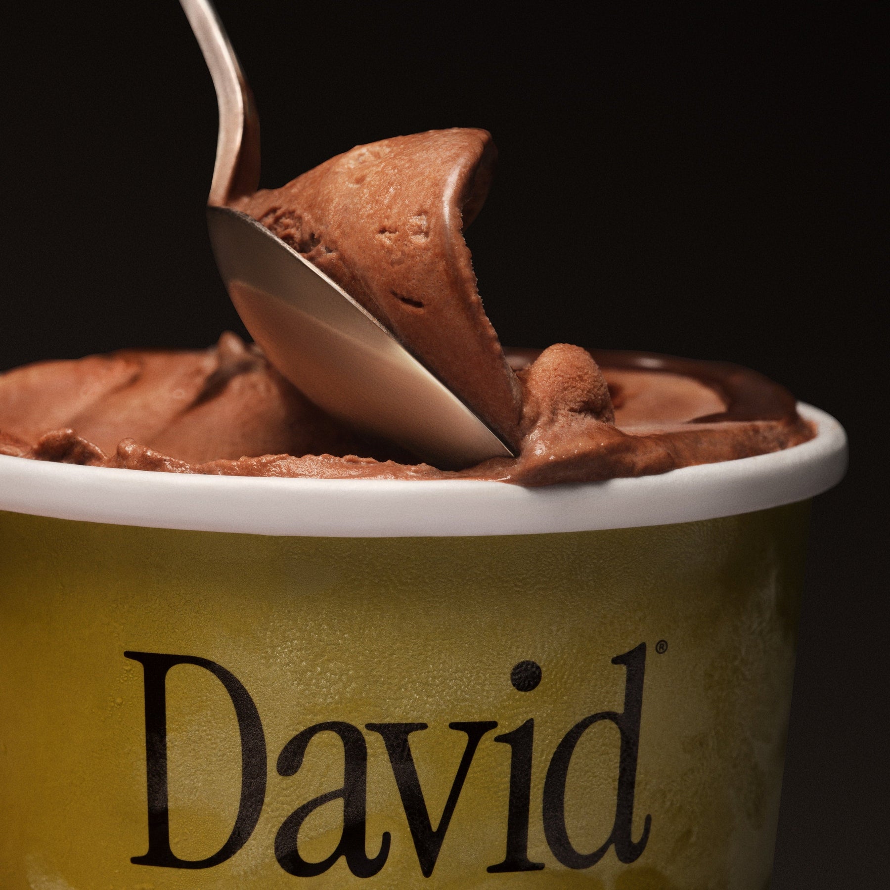 Spoon dipping into a container of chocolate ice cream branded 'David' on a black background