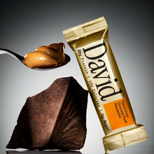David protein bar with a spoonful of peanut butter and a piece of chocolate on a gray background